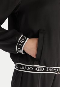 Giacca in satin nera con una vita elastica bianca e nera con logo "LIU-JO". Include una tasca laterale e polsini a costine.
