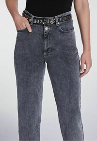 Jeans in denim grigio con vita alta e gamba dritta, dotati di una cintura nera con borchie e tasche frontali. Leggera scoloritura e dettagli di cucitura visibili.