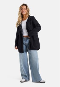 Veste noire à capuche avec poches, dotée d'une fermeture éclair à l'avant, d'accents dorés et d'une coupe décontractée. Associée à un jean large bleu clair.