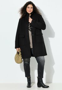 Cappotto di lana nero con colletto alto, due tasche frontali, abbinato a jeans grigi con borchie argentate e stivaletti neri. Borsa color marrone chiaro.