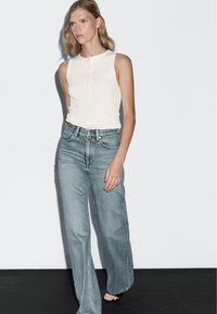 Top bianco senza maniche con bottoni frontali abbinato a jeans a gamba larga azzurri a vita alta. Texture liscia e design casual.