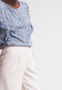 Blouse florale bleue avec épaules à volants, complétée par un pantalon crème sur mesure avec poches latérales et texture lisse.