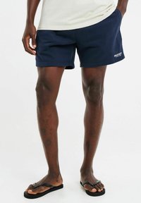 Navyblauwe zwembroek van zacht stof, met een platte tailleband en zijzakken. Logo gedrukt op de zoom. Slippers met dunne bandjes.