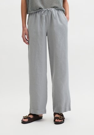 Losse lichtgrijze linnen broek met een trekkoord in de taille, gedragen met donkerbruine sandalen met gouden gespen en geschulpte randen.