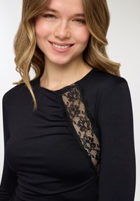 Top nero a maniche lunghe realizzato in tessuto strutturato con un pannello di pizzo laterale che mostra motivi floreali e bordi sfrangiati.