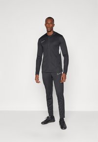 Schwarzer sportlicher Trainingsanzug, bestehend aus einer Zip-Jacke mit weißen Akzenten und passenden Hosen, aus glattem, leichtem Stoff gefertigt.