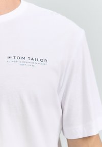 Vitt bomullströja med rund halsringning och kortärmar. Framsidan har marinblå text: "TOM TAILOR AUTHENTIC DENIM DEPARTMENT DEPT. (1962)".