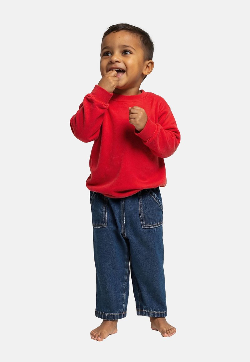 Sweat-shirt rouge à manches longues associé à un pantalon en denim bleu à jambe droite. L'enfant est debout pieds nus avec une expression joyeuse, la main dans la bouche.