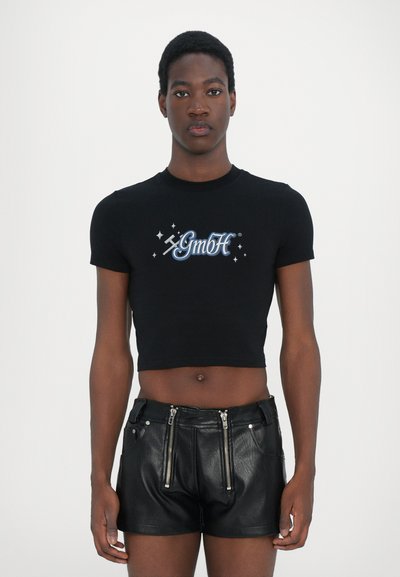 Black cropped t-shirt with "GmbH" logo in blue and white, paired with black leather shorts featuring zippers on the front pockets.