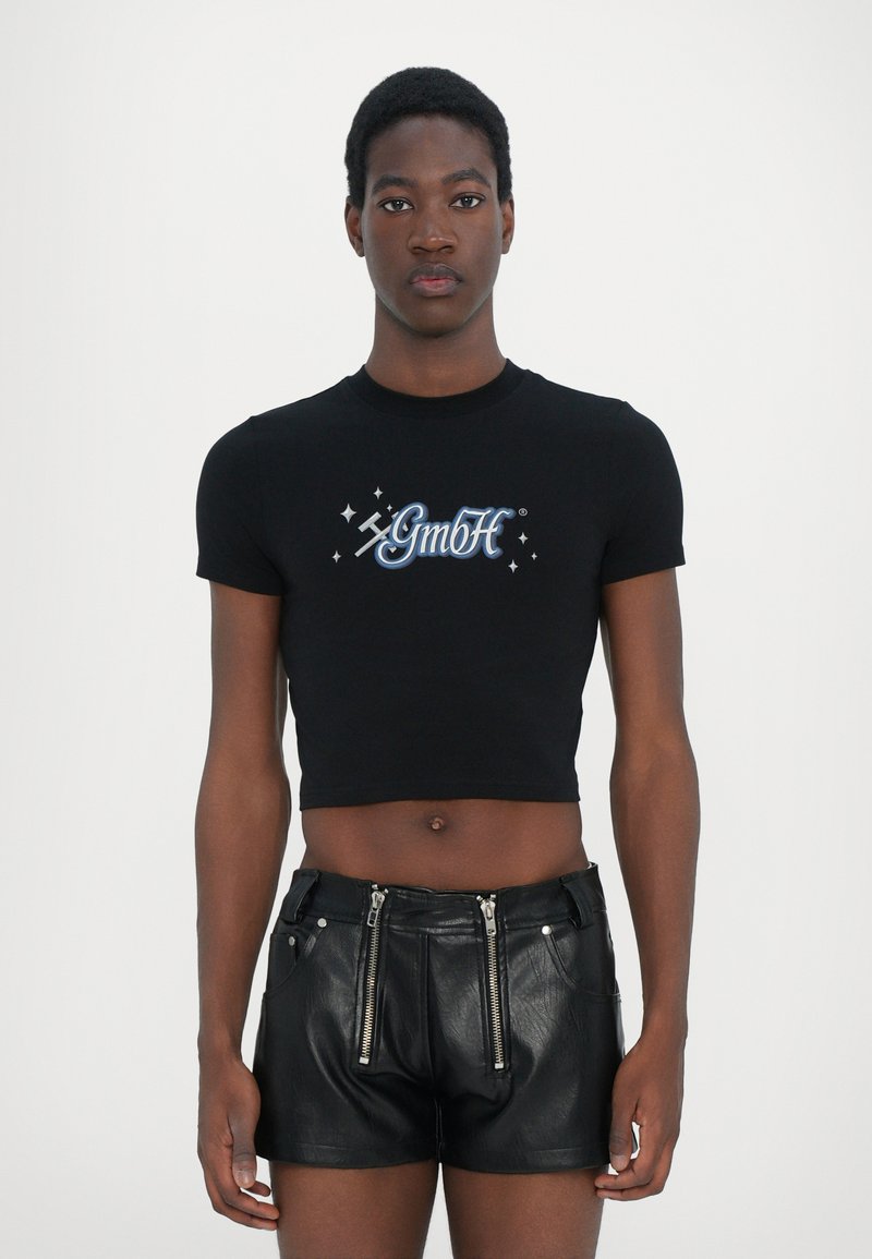 Tricou croppat negru cu logo-ul "GmbH" în albastru și alb, asortat cu pantaloni scurți din piele neagră cu fermoare la buzunarele din față.