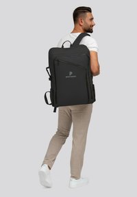 Sac à dos noir avec plusieurs compartiments, bretelles ajustables et un logo. Le tissu a une texture lisse, porté par une personne de dos.