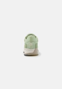 Sneaker verde chiaro con linguetta e suola bianca maculata vista da dietro su uno sfondo bianco.
