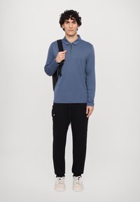 Blå langærmet polo-shirt med lynlås krave, parret med sorte cargo-bukser med sidelommer. Modellen bærer en sort rygsæk.