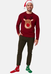 Roter Strickpullover mit Rentiermotiven, mit braunen und roten Akzenten. Olivgrüne Hosen und mehrfarbige festliche Socken runden das Outfit ab.