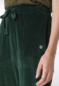 Samtgrüne Hose mit Kordelzugbund, seitlichen Taschen und einem kleinen Logoakzent am linken Bein. Glatte Textur mit weichem Finish.