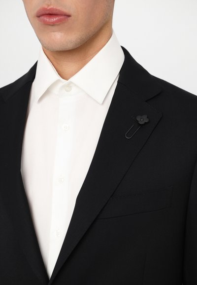 Chaqueta de traje negra ajustada con una camisa blanca debajo. Presenta un pin de solapa en forma de flor negra y un bolsillo en el lado izquierdo.