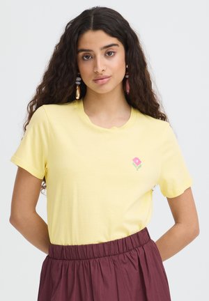 Noor naine, kellel on pikad lokkis juuksed, kannab kollast T-särki, millel on väike roosa lillekujundus, ning kõrge vöökohaga bordoopunast seelikut.