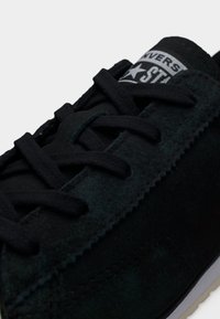 Κοντινό πλάνο μαύρου σουέντ αθλητικού παπουτσιού με μαύρα κορδόνια και ορατό λογότυπο "Converse Star" στη γλώσσα, πάνω σε λευκό φόντο.