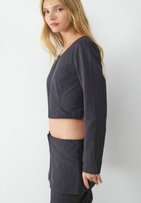 Gekürzter dunkelgrauer Blazer mit dunkelgrauem Nadelstreifen, langen Ärmeln und rundem Ausschnitt, kombiniert mit passenden Hosen, die über eine Seitenasche verfügen.