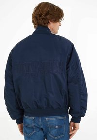 Giacca bomber blu navy realizzata in tessuto liscio con colletto a coste e polsini elasticizzati. Logo "TOMMY JEANS" ricamato sul retro.
