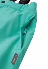 Pantalons imperméables turquoise avec taille ajustable, poche zippée sur le côté, et une étiquette indiquant "reima TEC". Texture de tissu lisse et durable.