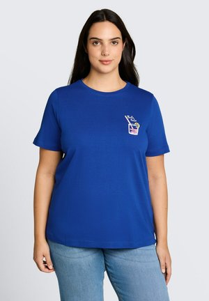 Kobieta nosząca niebieską koszulkę z krótkim rękawem z małą grafiką napoju "Bella Italia" na lewym piersi, w połączeniu z jasnoniebieskimi dżinsami.