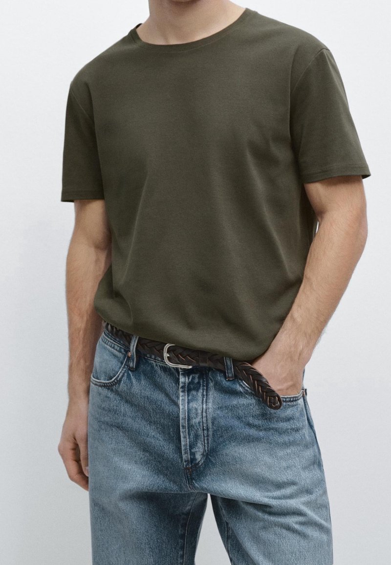 Homme portant un t-shirt à manches courtes vert olive rentré dans un jean bleu clair avec une ceinture tressée marron, une main dans la poche.