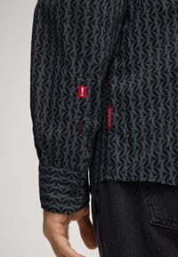 Camicia a maniche lunghe nera con motivo a zigzag, chiusure a bottone sui polsini e etichette rosse del marchio sulle maniche e sull'orlo inferiore.