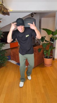 Μαύρο καπέλο με ανάγλυφο λογότυπο, μαύρο μπλουζάκι, παντελόνι ελιάς, λευκά αθλητικά παπούτσια, εσωτερικός χώρος με ξύλινο πάτωμα και φυτά εσωτερικού χώρου.