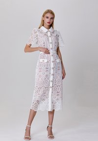Robe en dentelle blanche avec un col, des manches courtes, un devant boutonné et une ceinture assortie ; présente des motifs floraux et des poches plaquées.