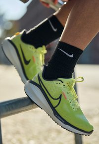 Lime roheline sportkingad tekstuuriga ülemise osaga, tume sinine aktsent ja silmapaistev must swooshi logo. Asetatud piirde peale.
