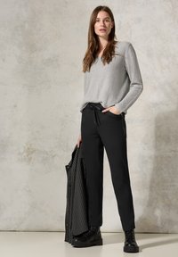 Gray, long-sleeve top made of soft fabric, paired with black drawstring pants featuring side zippers. Model holds a striped jacket. Black boots complete the look.