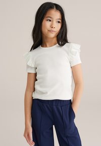 Top bianco a maniche corte con costine e dettagli a volant verde chiaro sulle spalle, abbinato a pantaloni blu navy, caratterizzati da una vestibilità comoda e tasche.