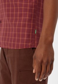Korte mouwen, kastanjebruin geruit shirt met oranje strepen. Heeft een groene merketiket en wordt gedragen met een bruine broek. Gestructureerde stof.