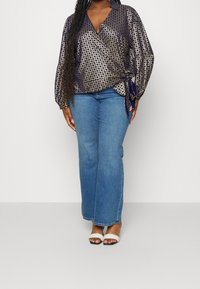 Top avvolgente blu navy con motivo geometrico dorato, maniche lunghe e dettaglio fiocco in vita, abbinato a jeans svasati azzurri e tacchi bianchi.