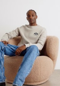 Beige sweatshirt med broderet logo, parret med lys blå denim jeans. Sidder på en rund, fuzzy, beige lænestol. Enkel og moderne design.