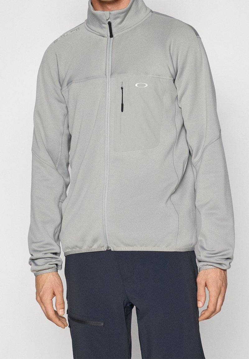 Homme portant une veste polaire grise claire zippée avec poche poitrine et pantalon noir avec poche zippée latérale, debout, bras détendus.