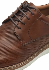 Scarpa in pelle marrone caratterizzata da una superficie tessurizzata con un design perforato, lacci grigi e suola bianca a contrasto.
