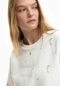 Mujer joven con cabello largo y rubio que lleva una sudadera blanca con el texto "DESIGUAL" y pequeñas decoraciones de cuentas florales en amarillo y verde.
