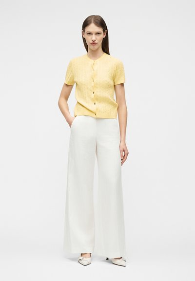 Γυναίκα που στέκεται φορώντας μια κίτρινη κοντομάνικη ζακέτα με κουμπιά, άσπρο παντελόνι φαρδιά μπατζάκια και λευκά μυτερά παπούτσια, μπροστά σε απλό φόντο.