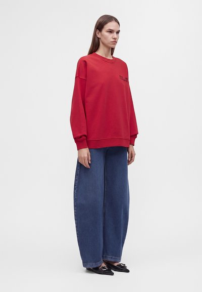 Femme aux longs cheveux bruns portant un sweat-shirt rouge oversize, un jean large bleu et des chaussures noires pointues, debout devant un fond uni.