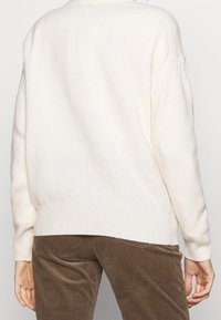 Pull en tricot de couleur crème, à coupe décontractée, avec des poignets et un ourlet côtelés. Porté avec un pantalon en velours côtelé marron foncé, mettant en valeur un contraste de textures.