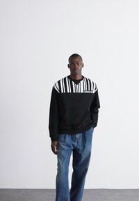 Sweatshirt preto com padrão de riscas brancas na parte superior, apresentando um decote redondo e o logo "Moschino". Usado com calças de ganga azuis.