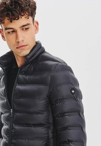 Czarna kurtka puchowa z wysokim kołnierzem, pikowanym wzorem i zamek błyskawiczny z przodu. Posiada naszywkę z logo na lewym ramieniu. Gładka, błyszcząca faktura.