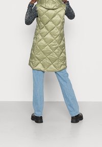 Pikowana zielona kamizelka z kapturem, o błyszczącej powierzchni, z bocznymi suwakami i dłuższym tyłem, w połączeniu z jasnoniebieskimi dżinsami i czarnymi butami.