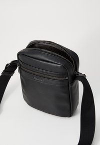 Schwarze Leder-Crossbody-Tasche mit strukturiertem Finish, doppelten Reißverschlussfächern und einem Riemen; mit Calvin Klein-Branding auf der Vorderseite.