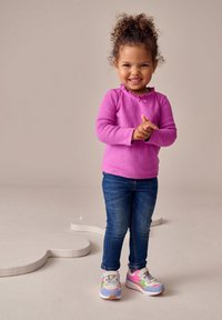 Top rosa de manga larga con cuello con volantes, jeans azules y zapatillas deportivas coloridas con un diseño de dibujos animados y detalles texturizados.