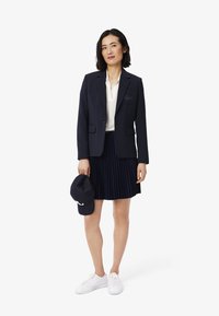 Marinblå kavaj med en knapp och bröstficka, vit blus, veckad marinblå kjol, marinblå mössa och vita sneakers. Släta texturer genomgående.