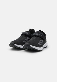 Schwarze Sportschuhe mit einem Textil-Obermaterial, ausgestattet mit drei seitlichen Streifen. Eine gepolsterte weiße Sohle und ein verstellbarer Riemen sorgen für einen sicheren Sitz.