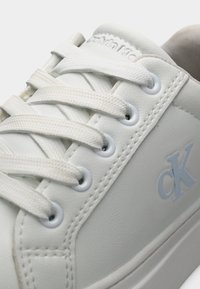 Λευκό δερμάτινο αθλητικό παπούτσι με υφασμένο επάνω μέρος, επίπεδα κορδόνια και ελαφρώς γαλάζιο λογότυπο "CK". Διαθέτει στρογγυλές τρύπες για τα κορδόνια και απαλό, μινιμαλιστικό σχέδιο.